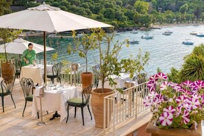 Un área de comedor al aire libre con mesas y sillas, con vista a un cuerpo de agua y embarcaciones.