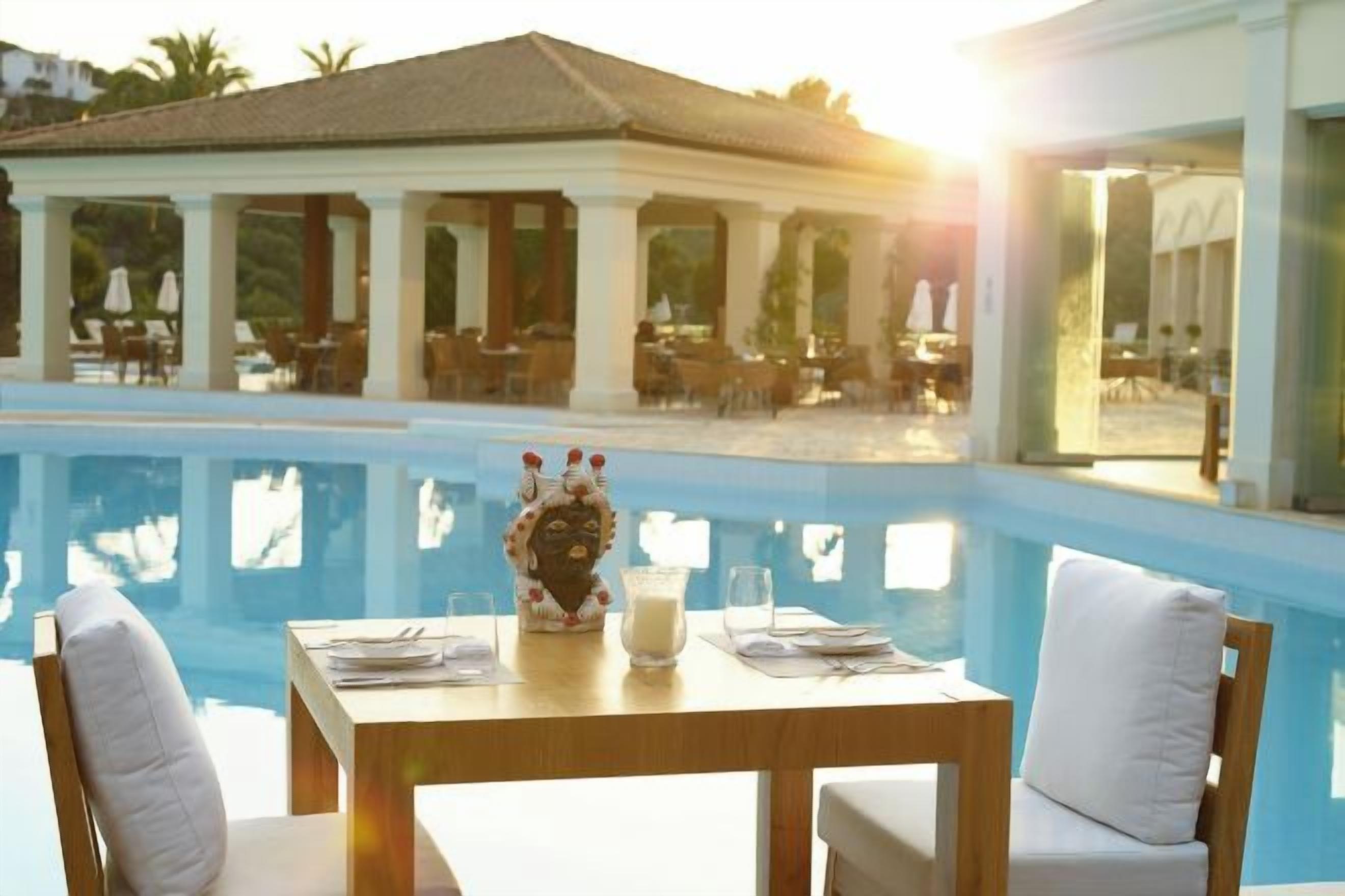 A dining table with chairs and a glass of water, overlooking a swimming pool with a building in the background.