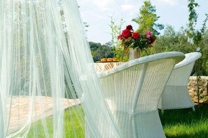 Una silla de mimbre con un dosel blanco, un florero con flores rojas y un muro de piedra al fondo.