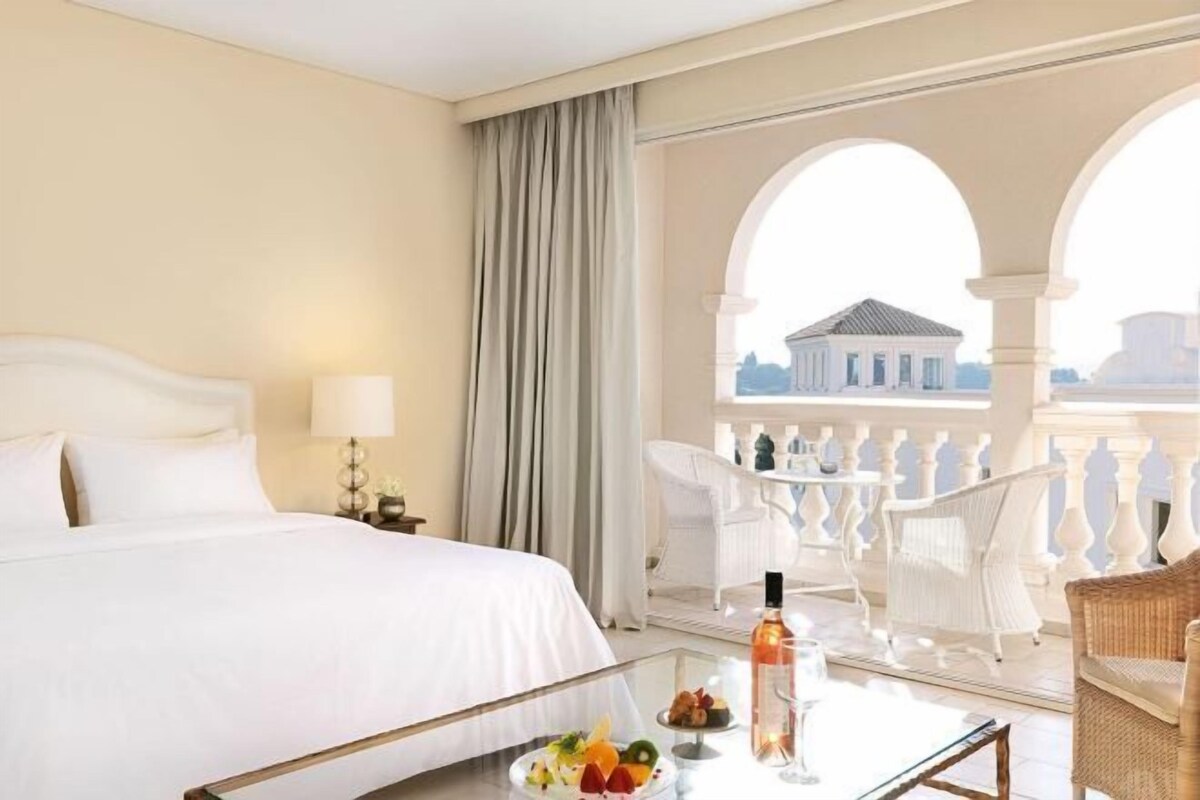 A hotel room with a bed, a glass coffee table with fruit and a bottle, white chairs, and a balcony with a view of a building.