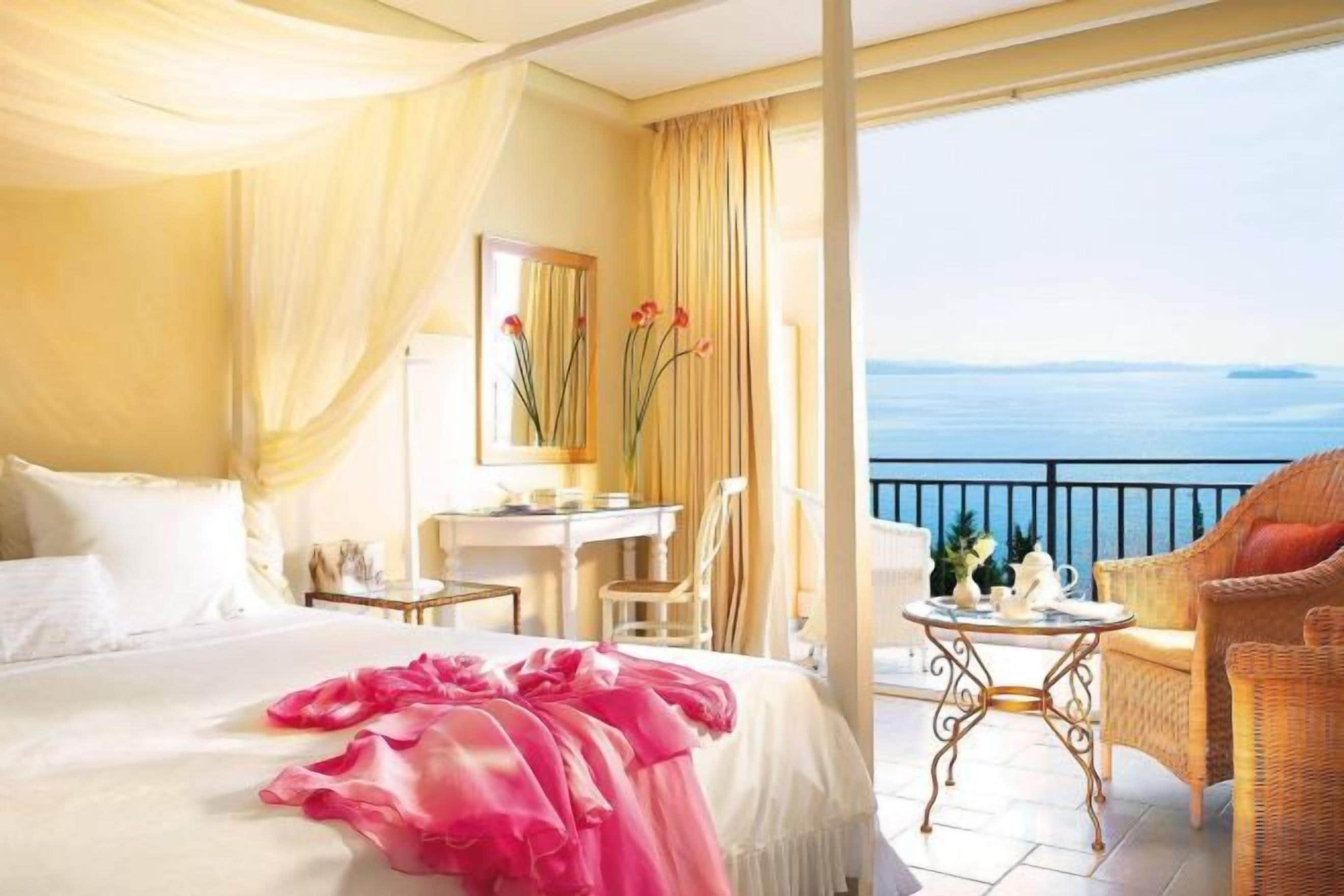 A hotel room with a bed, a desk, a chair, a small table, and a balcony with a view of the sea.