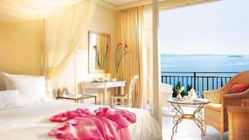 A hotel room with a bed, a desk, a chair, a small table, and a balcony with a view of the sea.