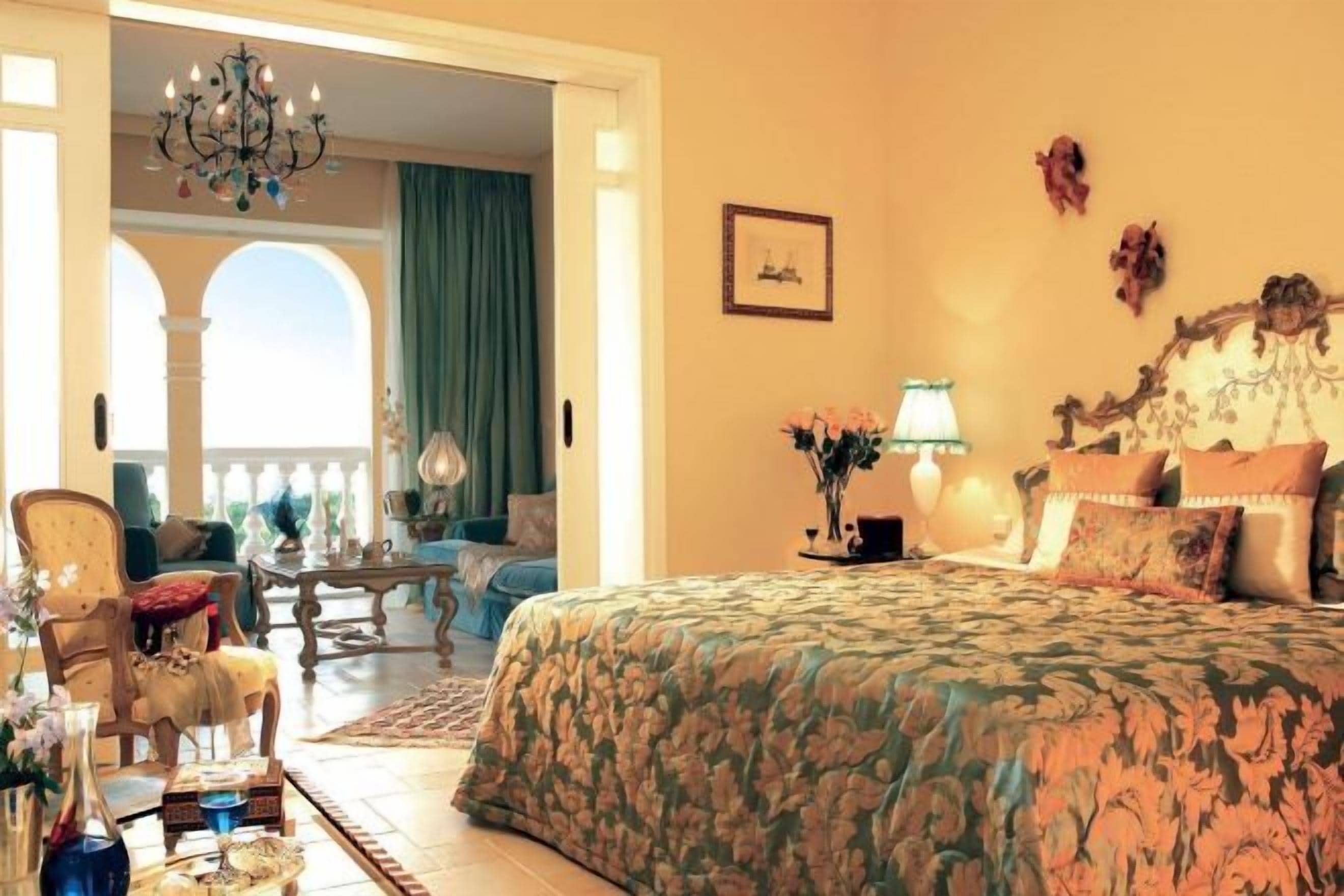 A bedroom with a large bed, a sitting area with a chair and a table, and a view of an outdoor balcony.