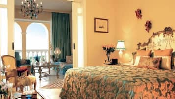 A bedroom with a large bed, a sitting area with a chair and a table, and a view of an outdoor balcony.