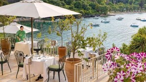 A terrace with tables and chairs, overlooking a body of water with boats. - Grecotel Eva Palace (Limni)