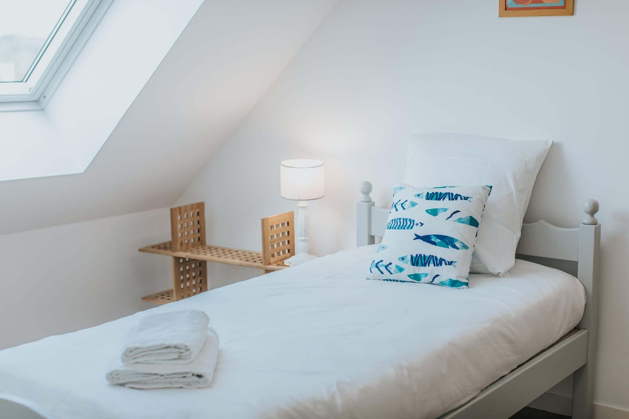 A neatly made bed with white bedding and a blue fish-patterned pillow, a bedside lamp, and a wooden chair.