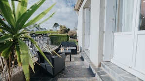 Une terrasse extérieure couverte, dotée d’un sol en pierre, de meubles en rotin noir et d’une plante en pot.