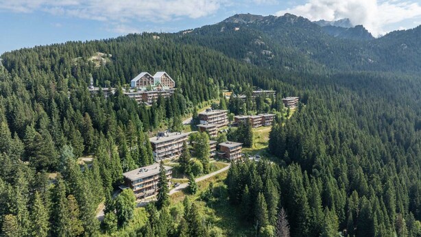 A resort with multiple buildings surrounded by dense forest, set against a backdrop of mountains. - Charming apartment on the ski slopes (Chamrousse)