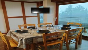A dining area with a table set for four, wooden chairs, a television mounted on the wall, and a view of trees through a large window. - Charming apartment on the ski slopes (Chamrousse)