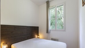 Une chambre à coucher avec une tête de lit en bois, un lit avec un matelas blanc, une lampe de chevet, une fenêtre avec des rideaux et un radiateur.