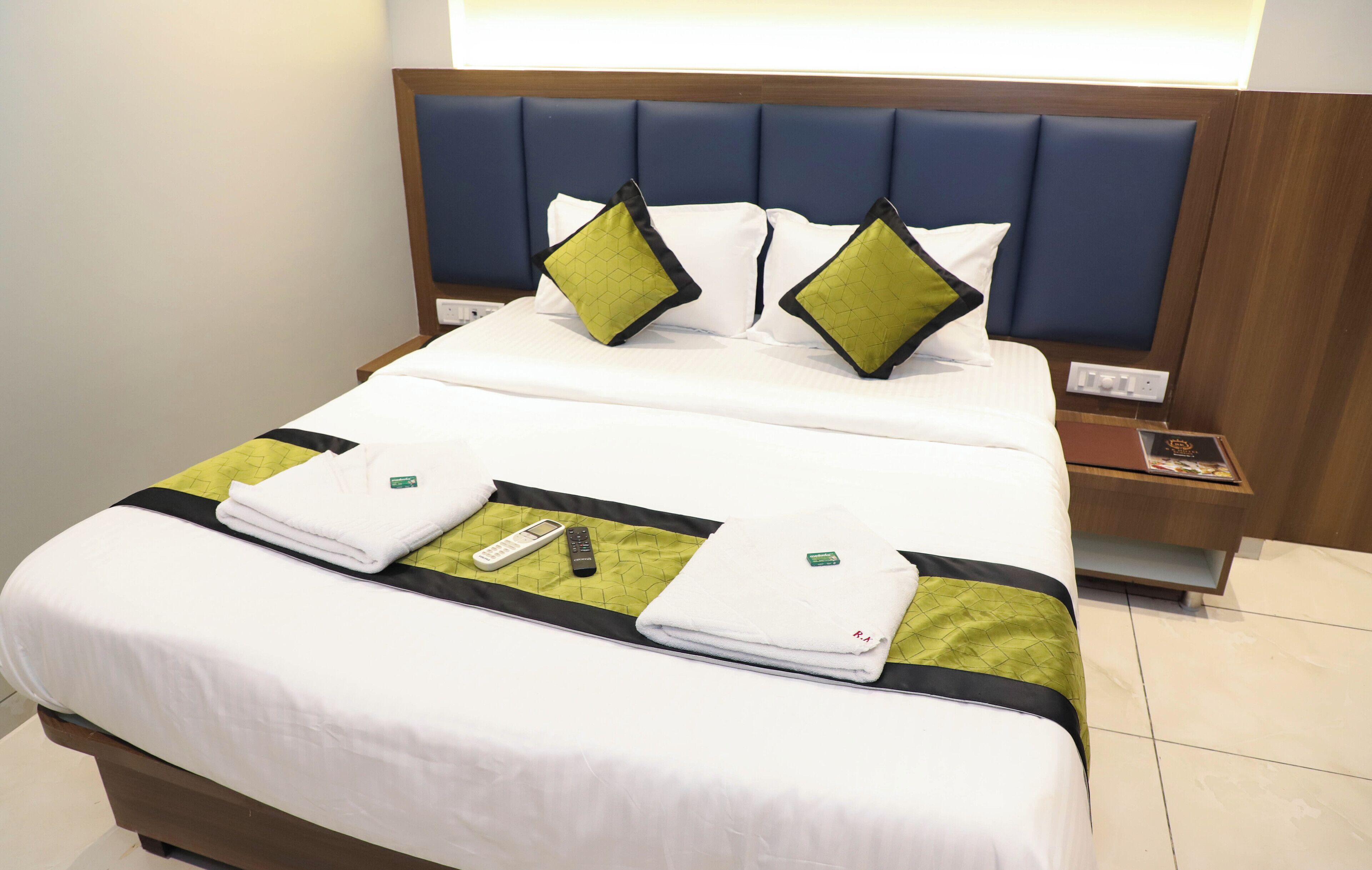 Una cama bien hecha con sábanas blancas, dos almohadas verdes y negras, un control remoto y una toalla doblada sobre un edredón verde.