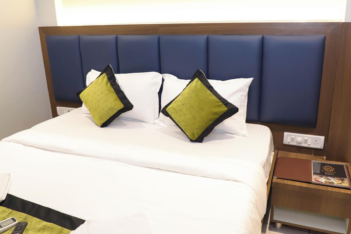 A hotel room with a neatly made bed, two green and black pillows, a white comforter, a wooden headboard, and a nightstand with a remote control and a menu.