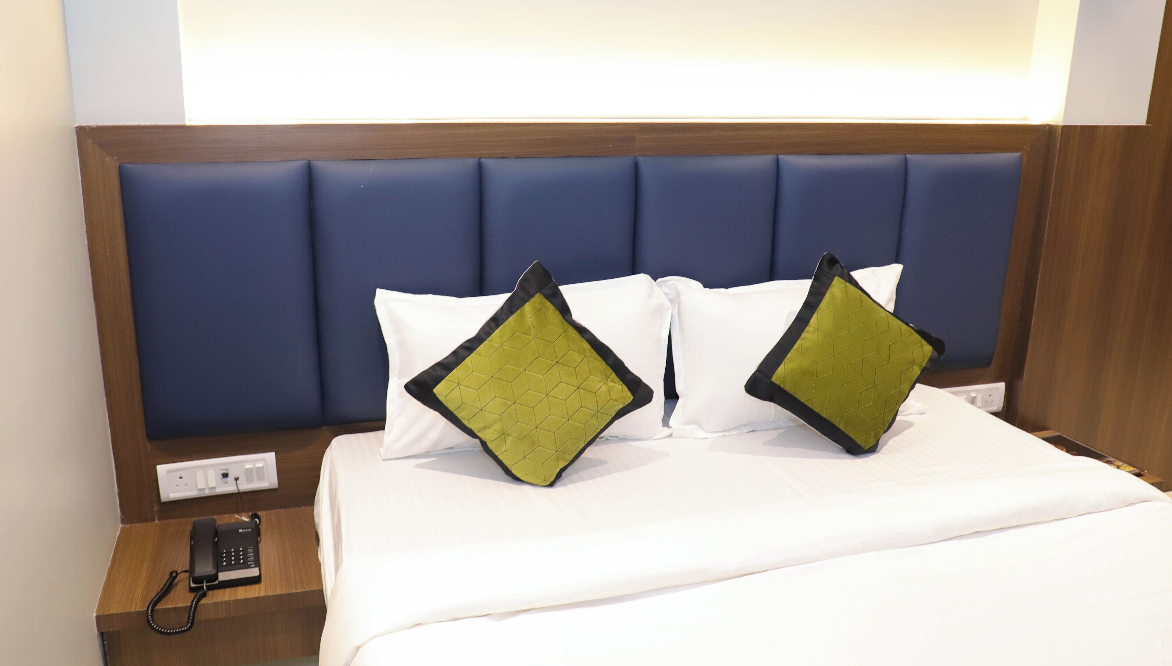 Une chambre d’hôtel avec une tête de lit en bois, deux oreillers blancs, deux coussins décoratifs verts et noirs, une couvre-lit blanche et un téléphone sur la table de chevet.