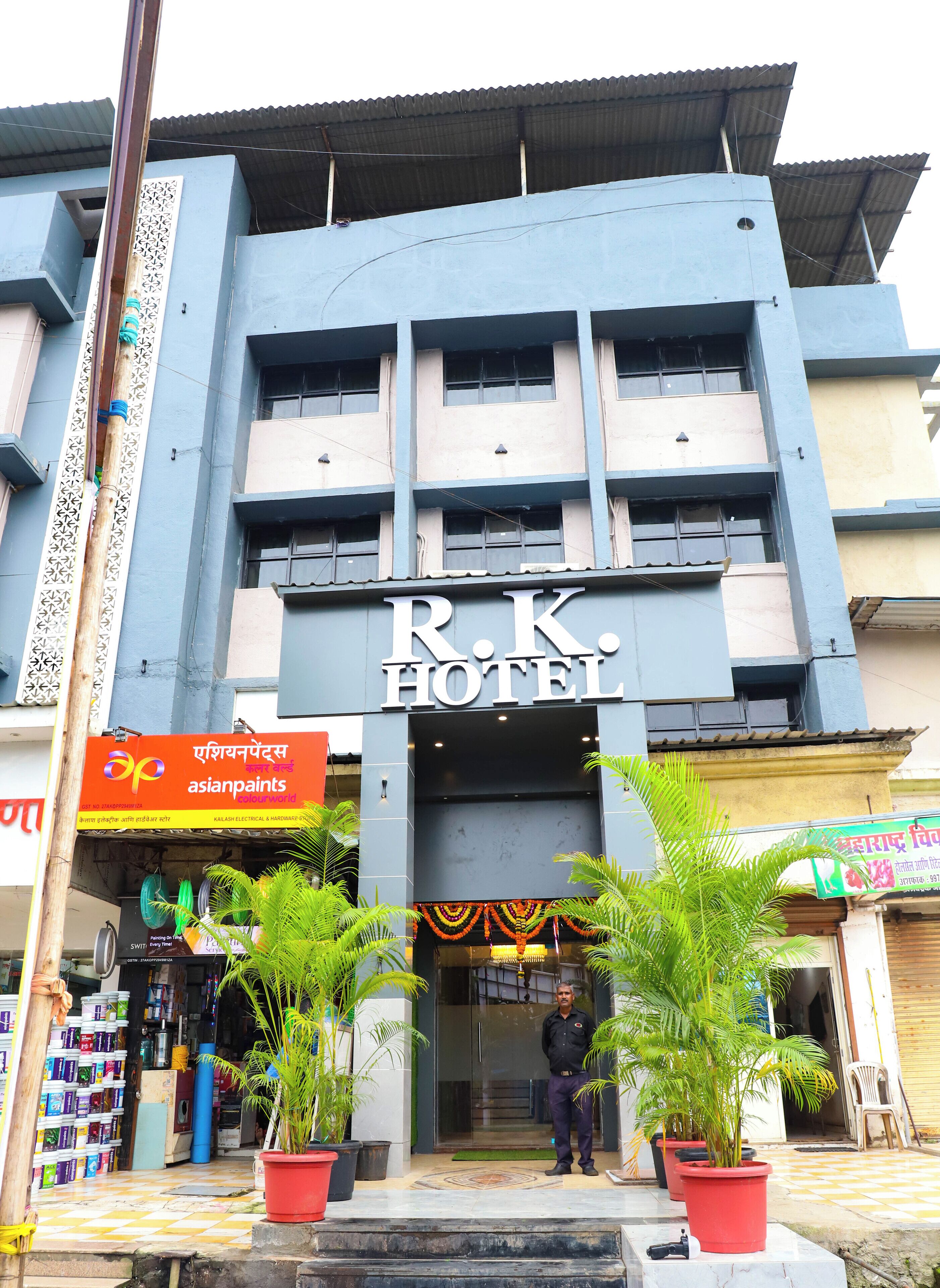 The exterior of R.K. Hotel with a signboard, a man standing at the entrance, and potted plants in front.