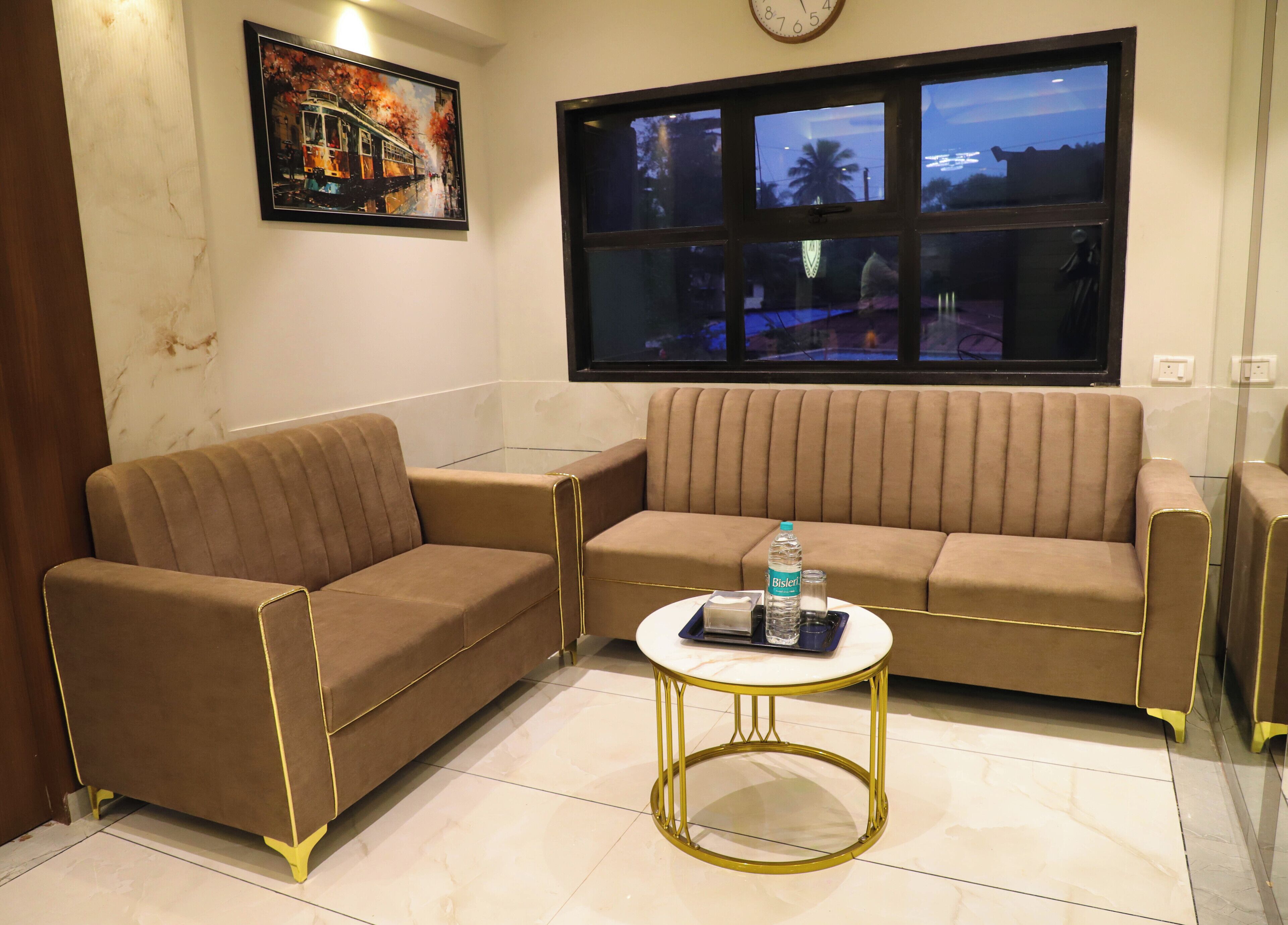 A room with a brown sofa, a small round table with a bottle and glasses, a clock on the wall, and a window with a view of palm trees and a sunset.