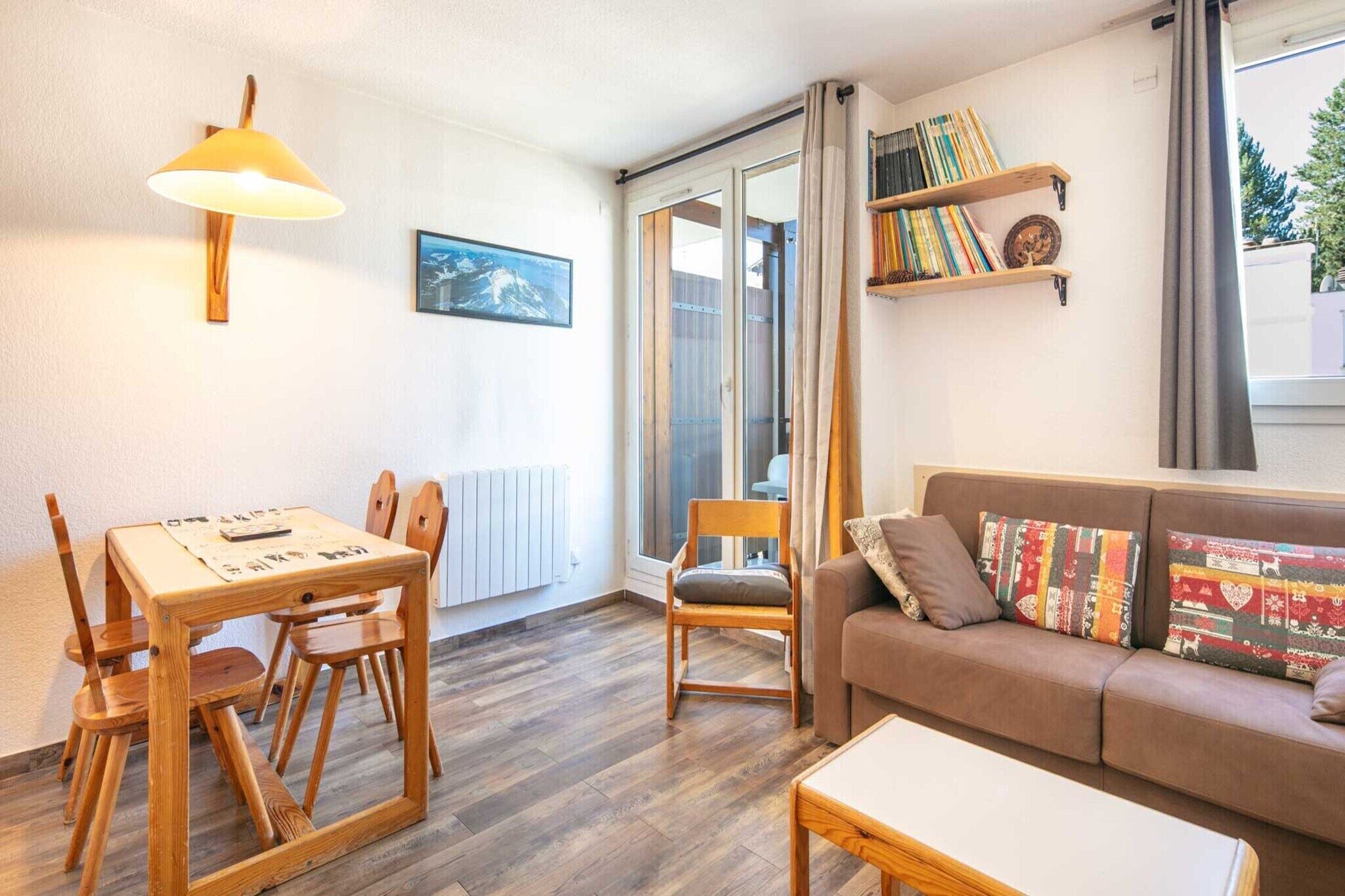A cozy living room with a wooden table, chairs, a sofa with patterned cushions, a bookshelf, and a large window with curtains.