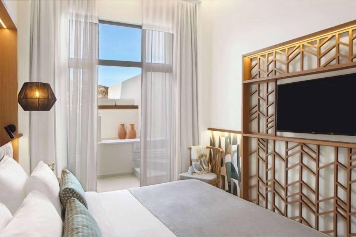 A modern hotel room with a large bed, a TV mounted on a wooden panel, and a view of the outdoors through a glass door.