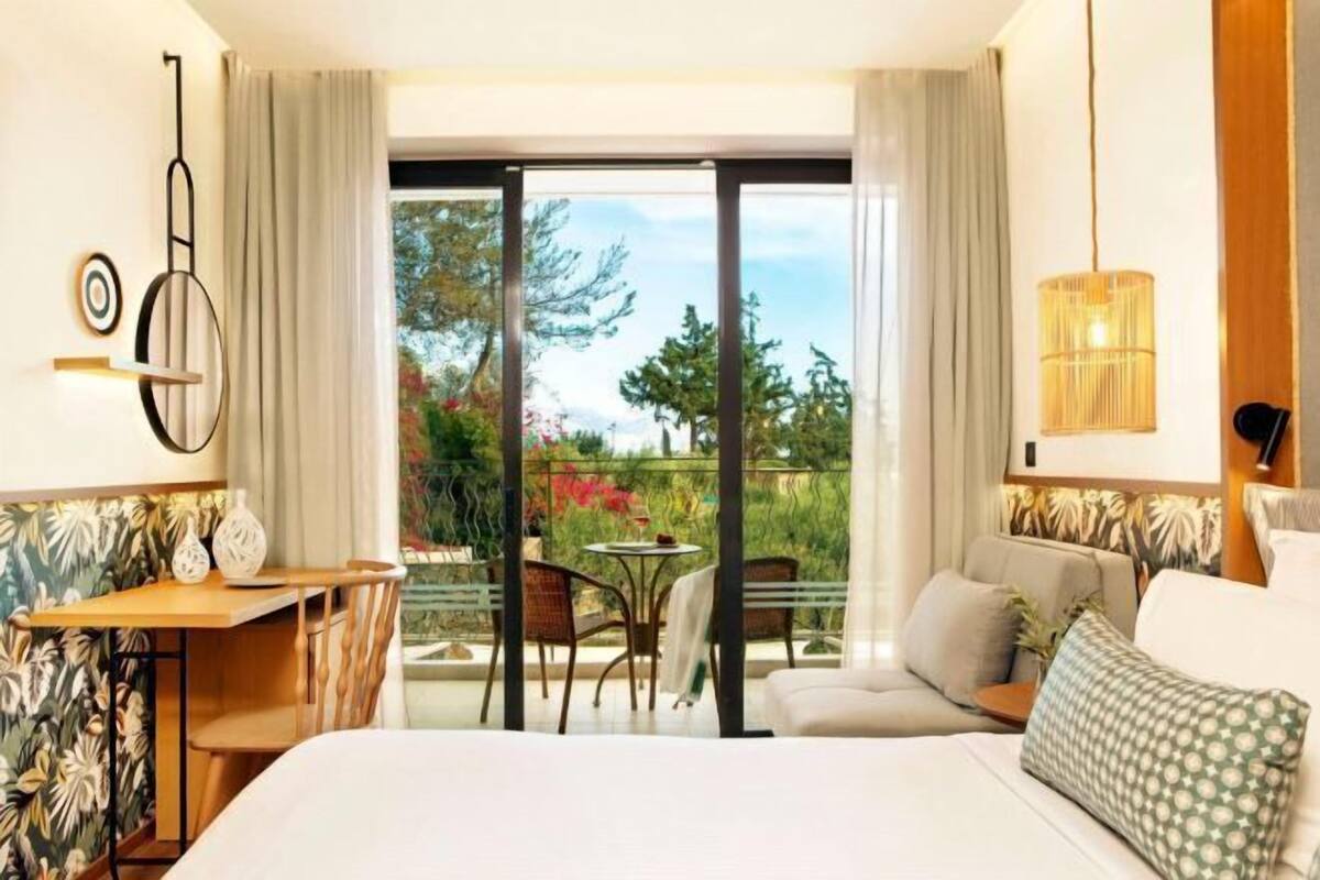 A hotel room with a bed, a desk, a chair, and a balcony with a view of greenery.