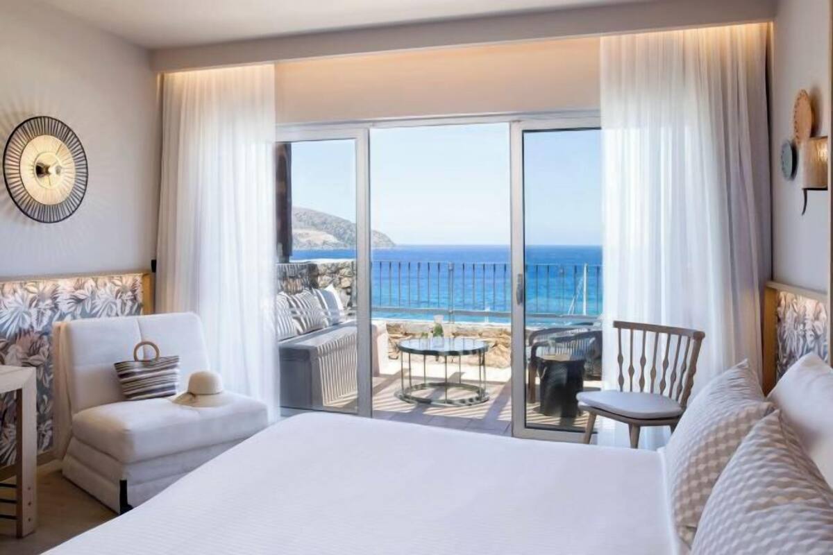 A hotel room with a large bed, a sofa, a chair, and a table. There is a balcony with a view of the sea.