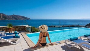 Un espace aménagé au bord de la piscine avec des chaises longues, une personne en maillot de bain et portant un chapeau, et une vue sur la mer et les montagnes.