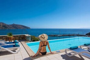 A poolside area with lounge chairs, a person in a swimsuit and hat, and a view of the sea and mountains. - Wyndham Grand Crete Mirabello Bay (Agios Nikolaos)