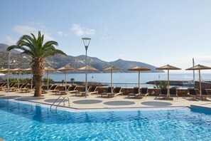 A swimming pool with lounge chairs and umbrellas, a palm tree, and a view of the sea and mountains. - Wyndham Grand Crete Mirabello Bay (Agios Nikolaos)