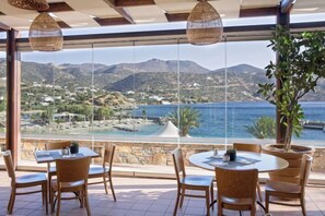A terrace with tables and chairs overlooking a scenic view of a coastal area. - Wyndham Grand Crete Mirabello Bay (Agios Nikolaos)