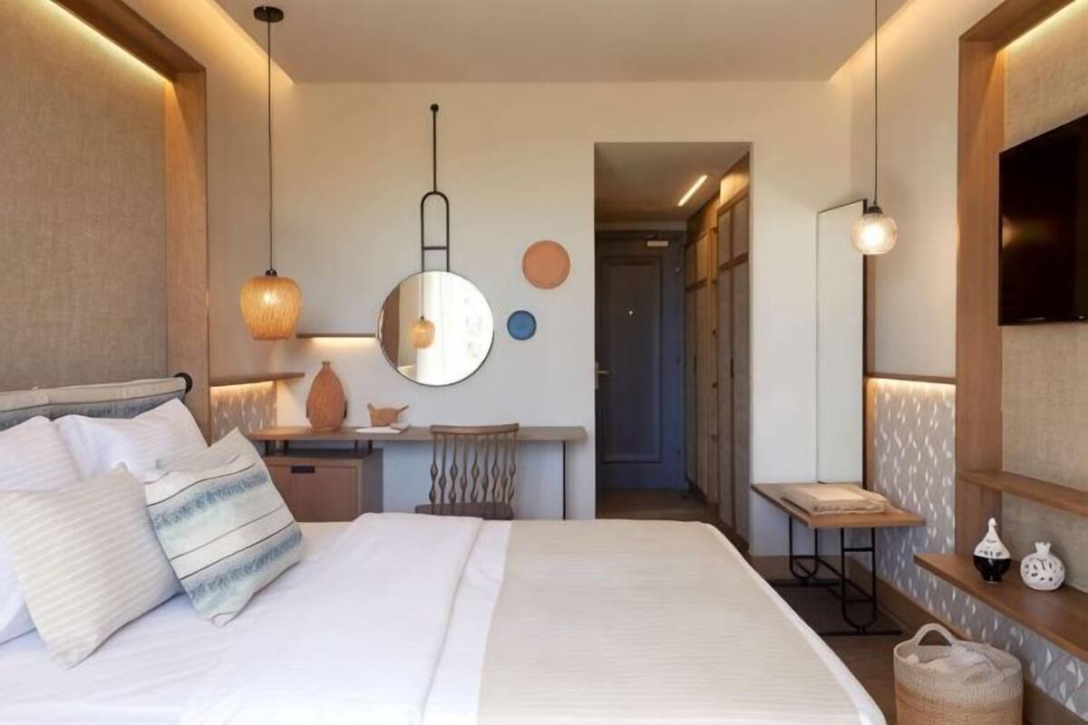 A bedroom with a bed, bedside table, lamp, mirror, and a view of a hallway.