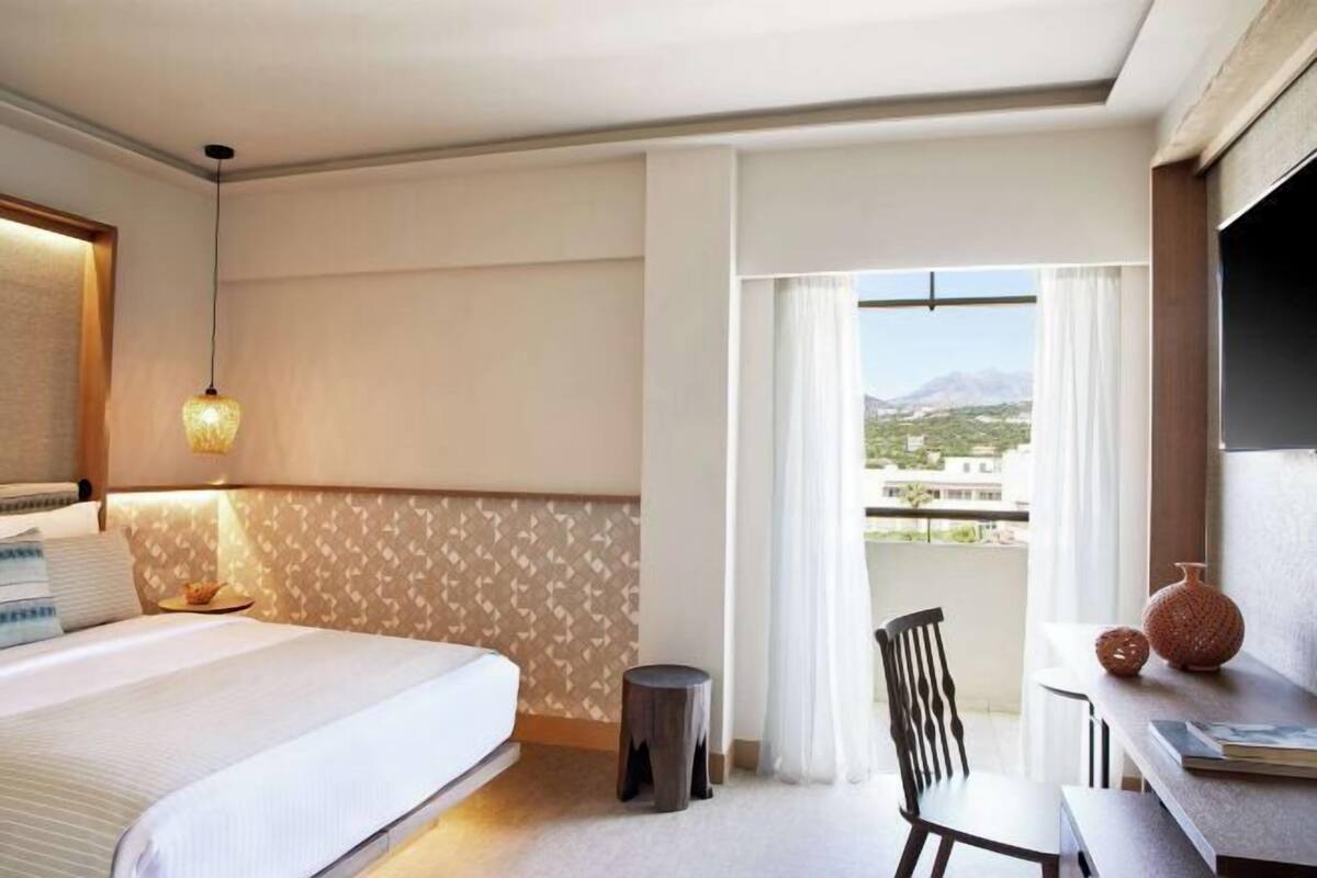 A hotel room with a bed, a small table, a chair, a lamp, and a view of the outdoors.