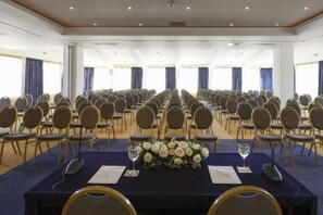 A conference room with rows of chairs, a long table with a floral centerpiece, and large windows with curtains. - Wyndham Grand Crete Mirabello Bay (Agios Nikolaos)