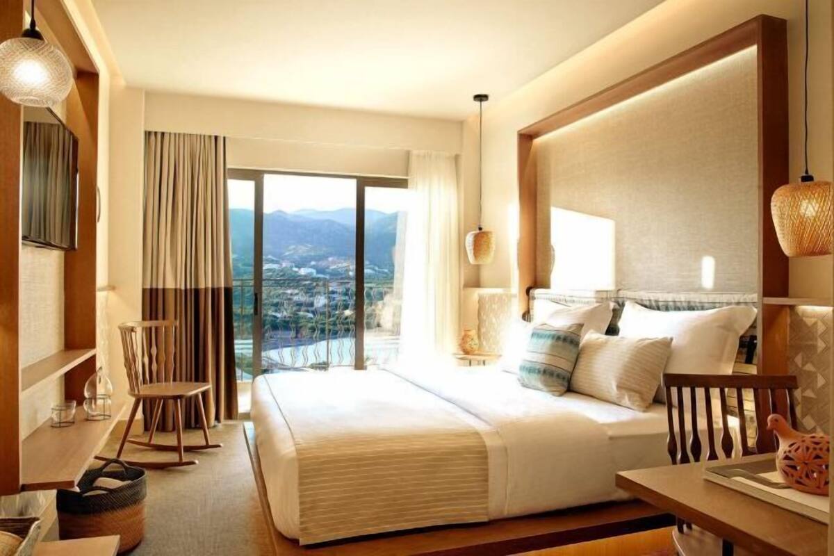 A hotel room with a large bed, a desk, a chair, a lamp, and a view of a cityscape.