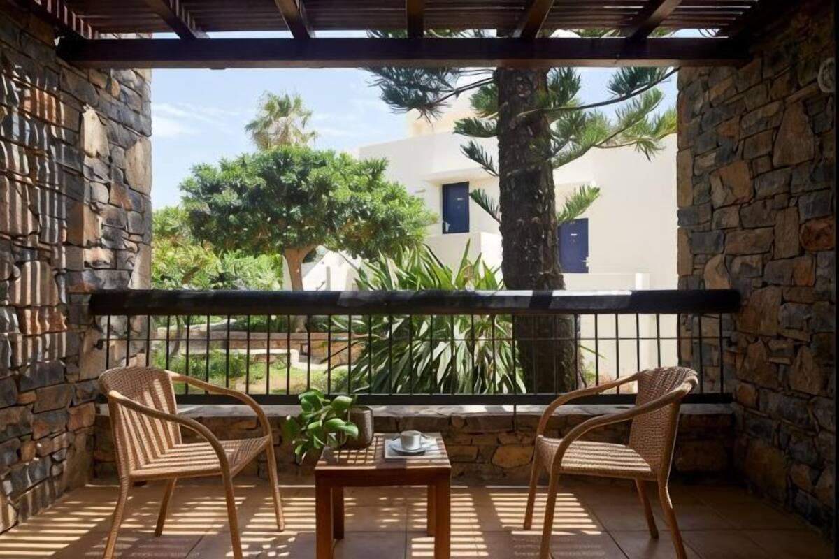A balcony with stone walls, two wicker chairs, a small table with a plant, and a view of a building and trees.