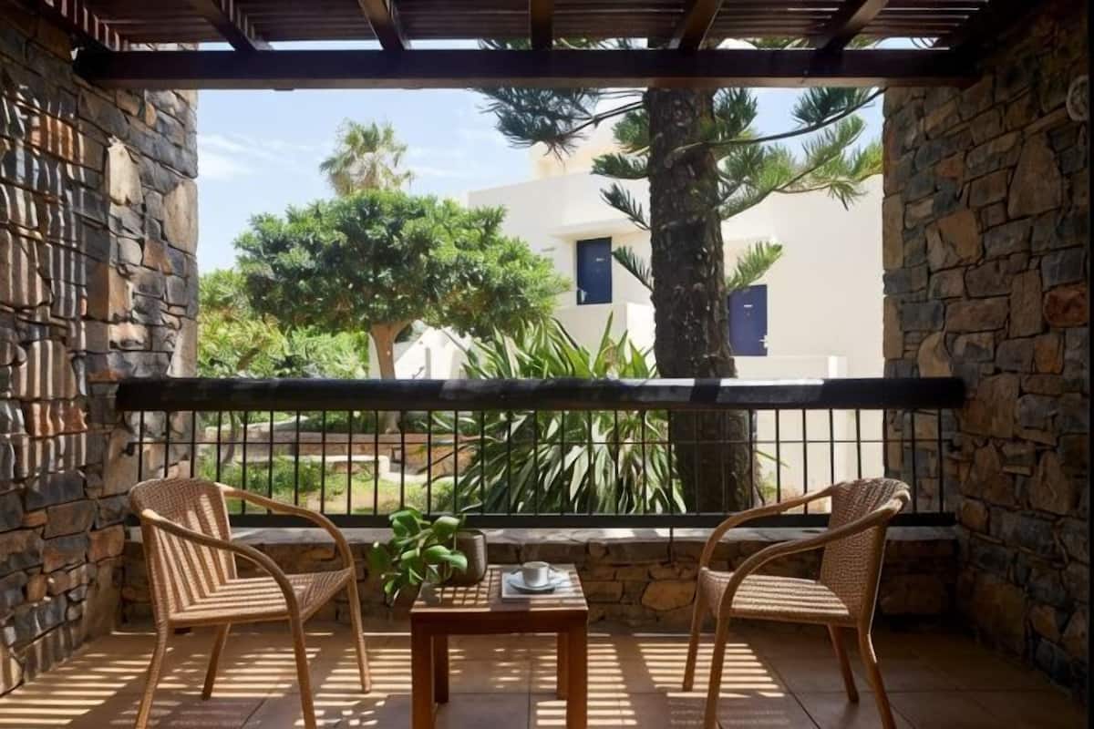 A balcony with stone walls, two wicker chairs, a small table with a plant, and a view of a building and trees.