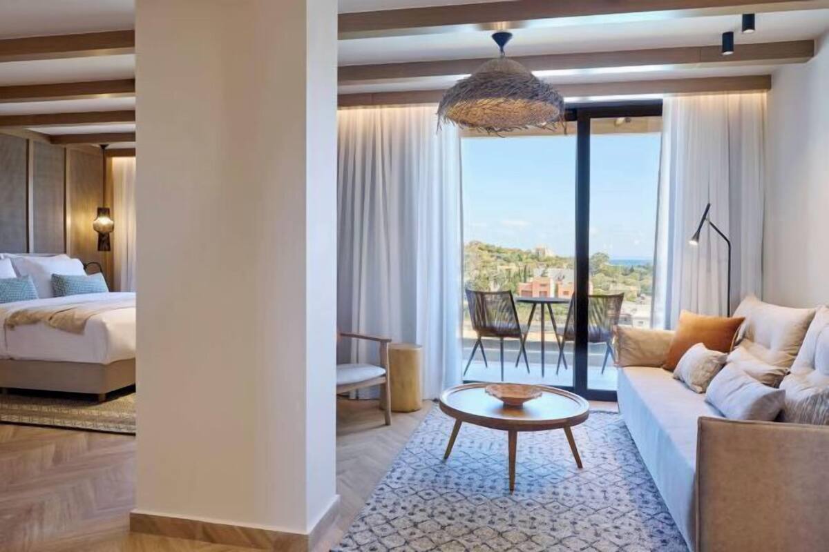 A modern hotel room with a bed, a sofa, a dining table, and a balcony with a view.