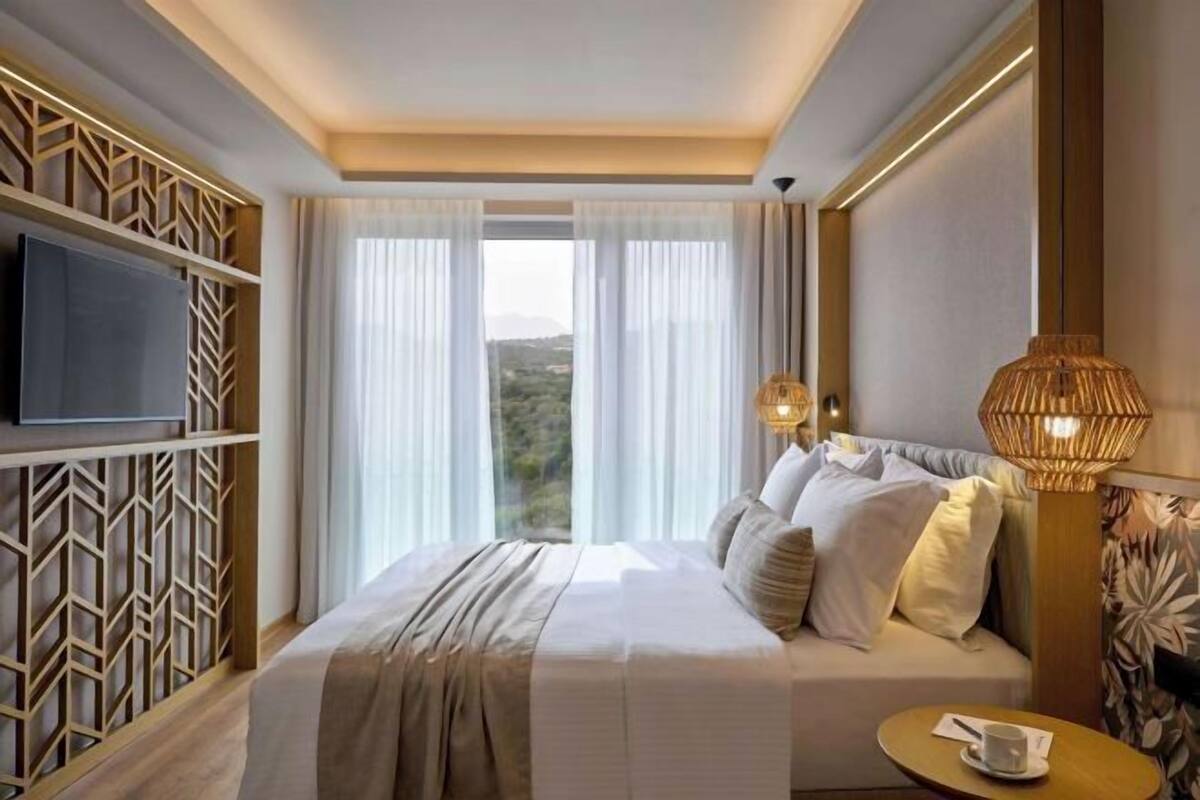 A hotel room with a bed, a TV, a window with curtains, a lamp, and a view of the outdoors.