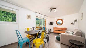 A dining area with a table and chairs, a sofa, a TV, and a window with a forest view. - Family-friendly apartment with terrace (Allos)