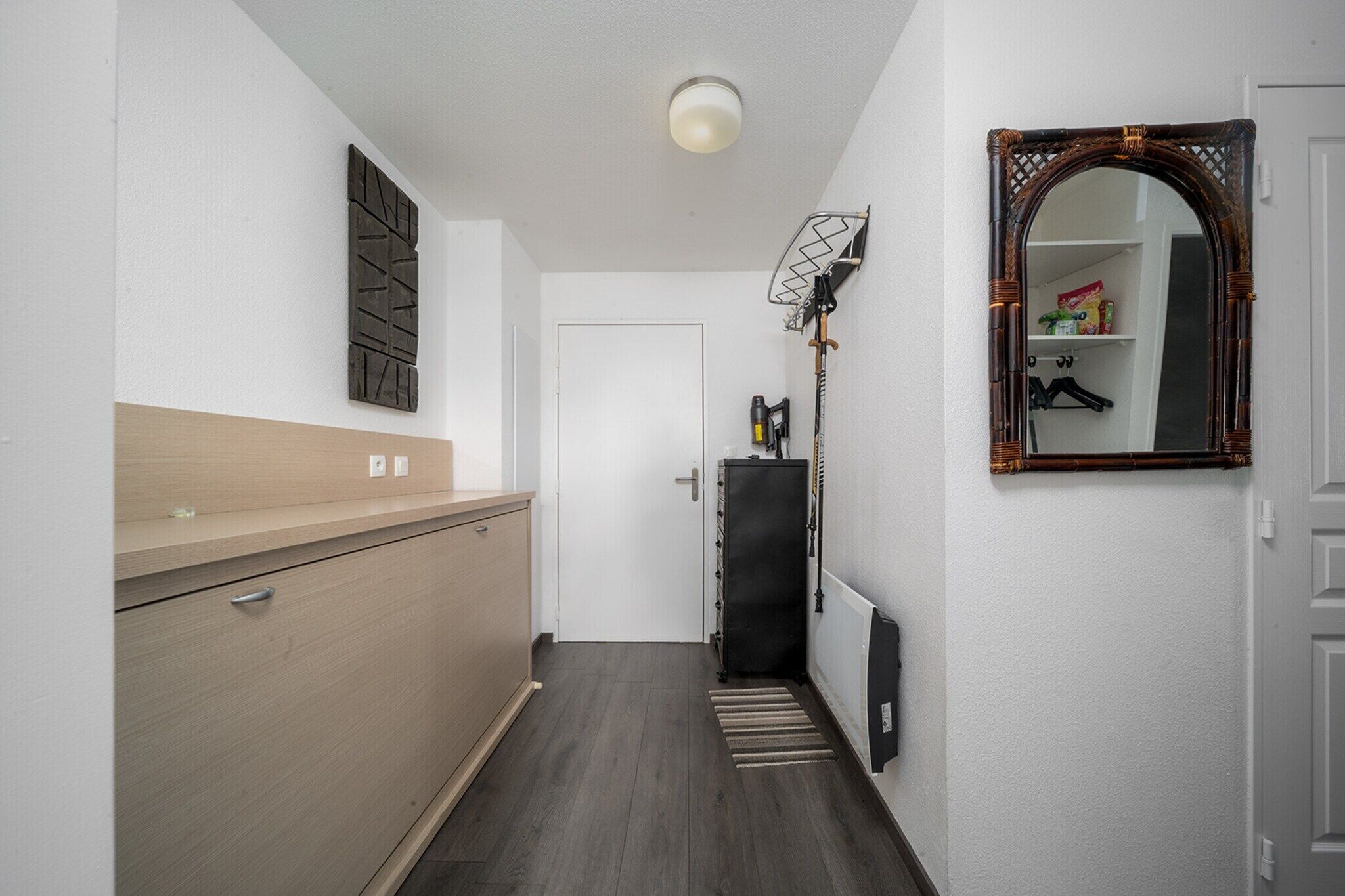 A narrow hallway with a wooden cabinet, a radiator, a mirror, and a door leading to a room.