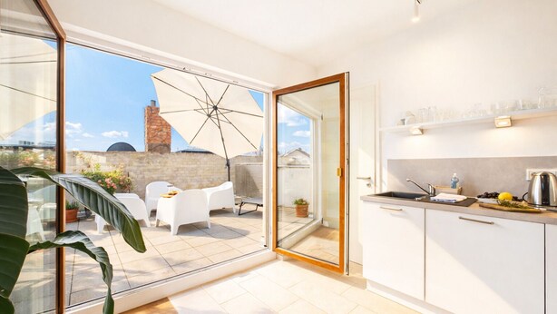 A modern kitchen with white cabinets, a sink, and a countertop with various items. There is a sliding glass door leading to an outdoor patio with white furniture and a large umbrella. - Eltville: The Eight – Urban Living (Eltville)