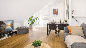 A modern living room with a flat-screen TV, a grey sofa, a dining table set for four, and a staircase leading to an upper level. - Eltville: The Eight – Urban Living (Eltville)