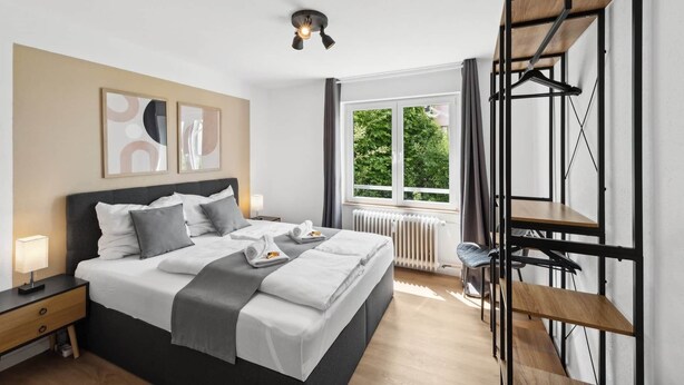 A modern bedroom with a bed, bedside tables, a radiator, a window with a view of greenery, and a black metal structure.