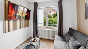 A modern living room with a grey sofa, a flat-screen TV mounted on a wooden panel, a round side table with a vase, and a window with curtains.
