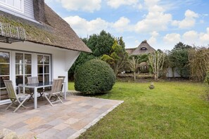 Une maison avec un toit de chaume, une terrasse avec une table et des chaises, et un jardin bien entretenu avec un grand arbuste et un mur de pierre en arrière-plan.