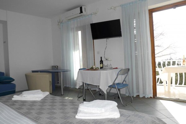 A hotel room with a bed, a desk, a chair, a table with a bottle and glasses, and a TV on the wall.