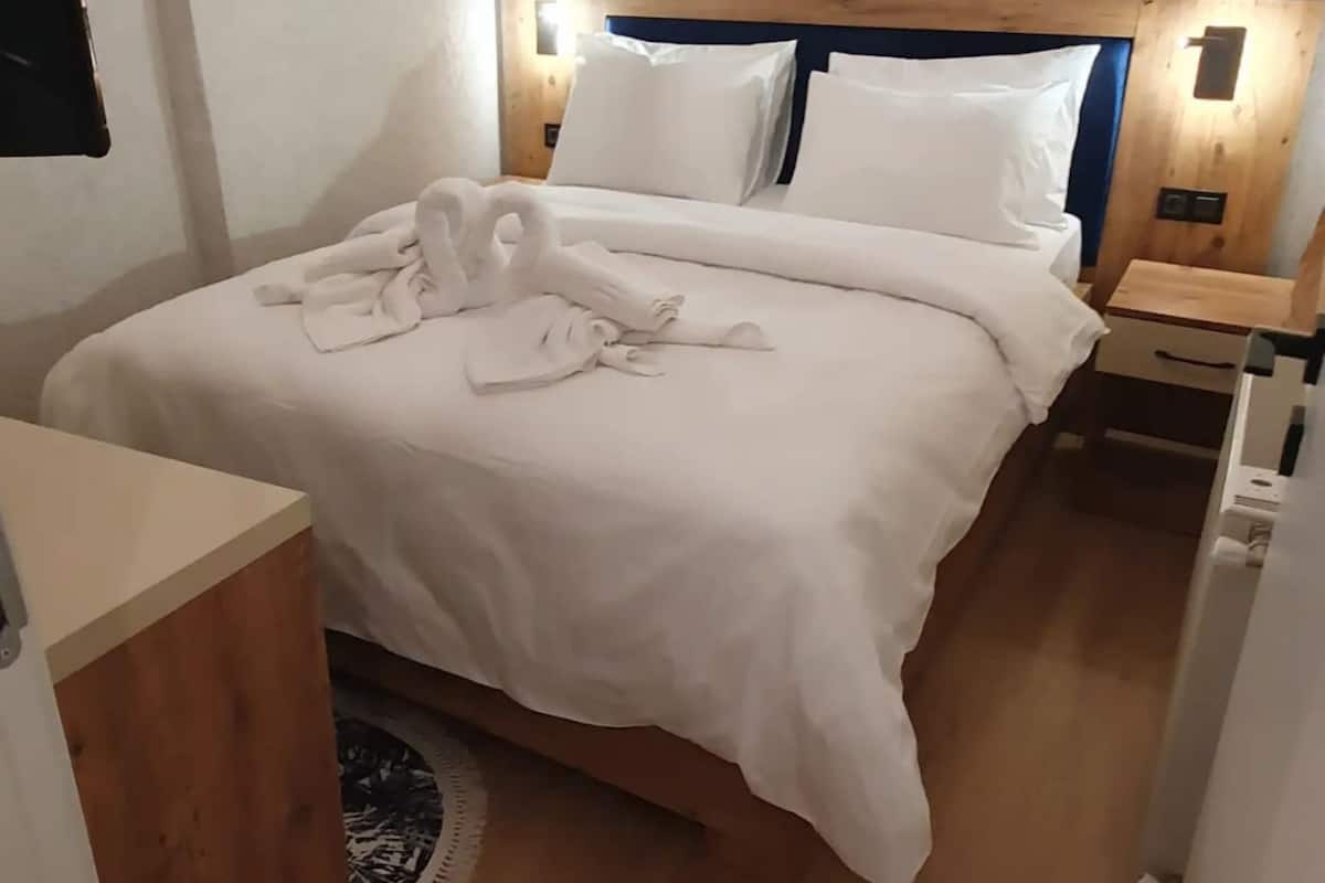 A neatly made bed with white linens and two pillows, a wooden headboard, a bedside table with a lamp, and a small round rug on the floor.