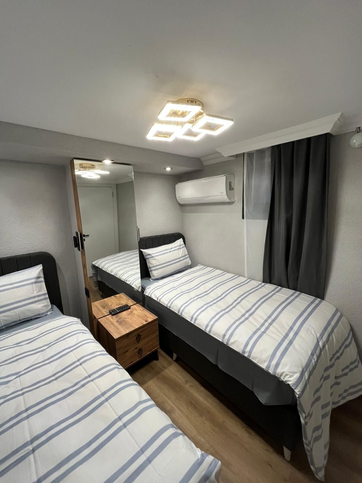 Una habitación pequeña con dos camas, un espejo, una mesita de noche y una lámpara en el techo.