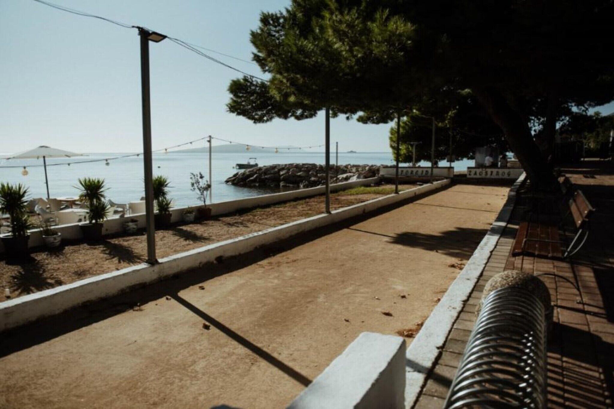 Une promenade dallée avec une rambarde en métal, un arbre et une vue sur la mer, avec un pont visible au loin.
