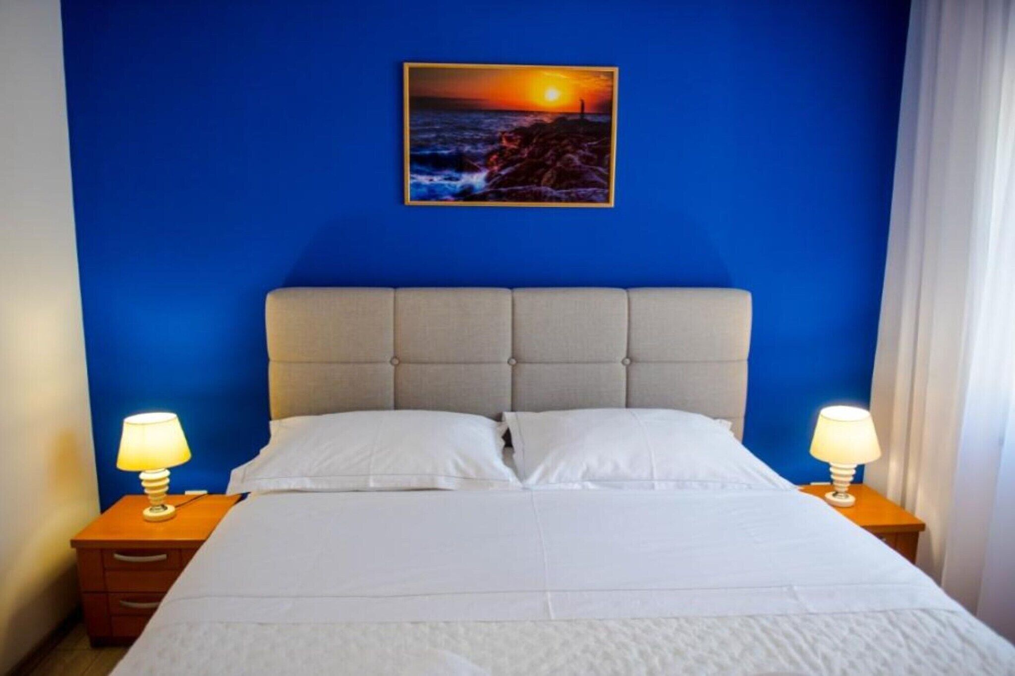 Ett hotellrum med en stor säng, två sängbord med lampor, en blå vägg och en tavla som föreställer en solnedgång.