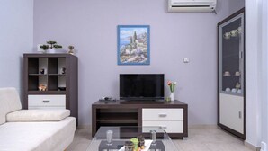 A modern living room with a sofa, a TV on a wooden stand, a glass coffee table, and a cabinet with decorative items. - One Bedroom Apartment with Balcony and Sea View 2 (Vrboska)