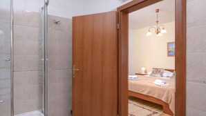 A bathroom with a glass shower enclosure, a wooden door leading to a bedroom with a bed, a nightstand, and a wall-mounted light fixture. - One Bedroom Apartment with Balcony 3 (Vrboska)