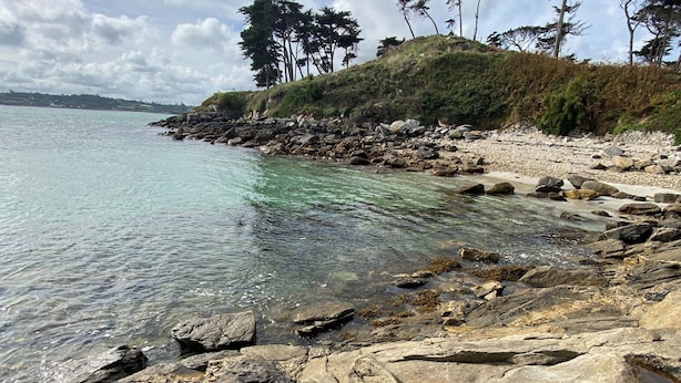 Un rivage rocheux bordé d’une eau claire, une personne assise sur les rochers et des arbres en arrière-plan.
